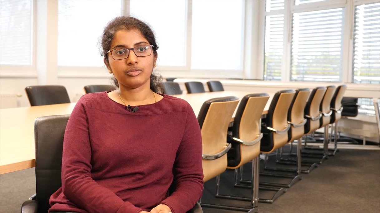 The picture shows Michelle Michael, an IT security consultant, sitting in a modern conference room. She is wearing a dark red top and glasses, her facial expression is focused and professional as she looks slightly past the camera. The background shows a large conference table with several empty chairs and large windows with slats that allow plenty of daylight into the room. This thumbnail is part of an employee testimonial and emphasizes Maxi Mustermann's expertise and commitment to IT security.